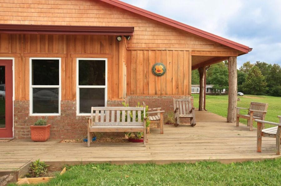 A wooden and brick house with a deck containing benches and rocking chairs sits on a grass field.