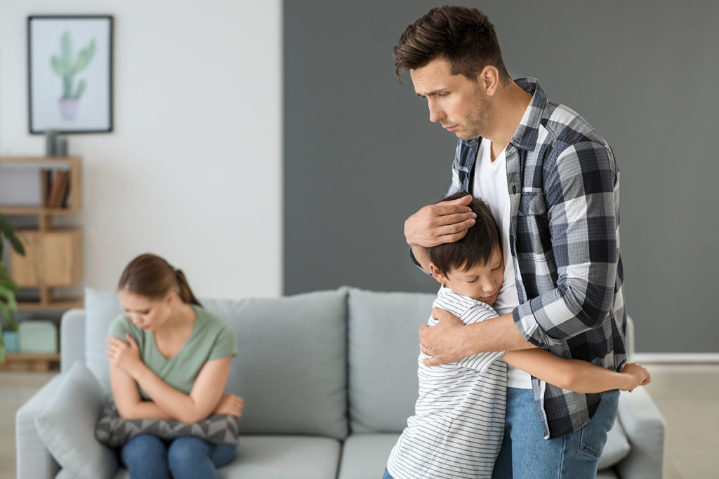child hugging worried father as mother sits on couch distraught about how addiction affects families