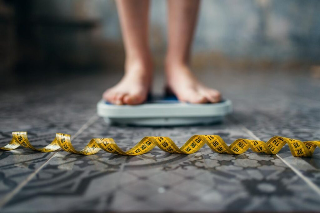 Person standing on scale with measuring tape in front of it considering starting a 12 step program for eating disorders