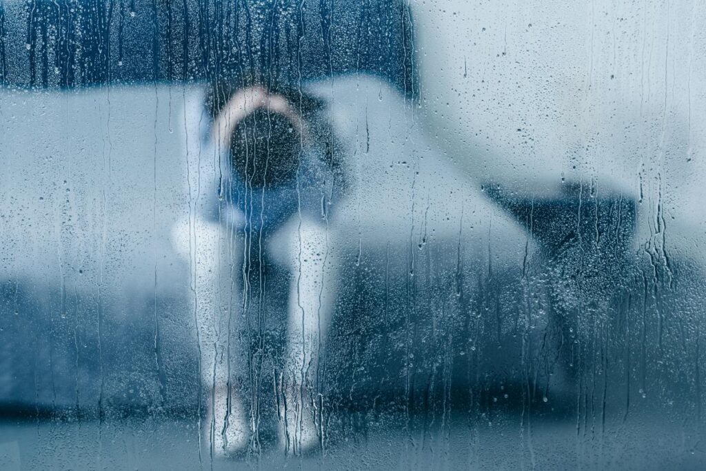 person sitting on edge of bed hanging head in despair on a rainy day after learning about how does weather impact addiction