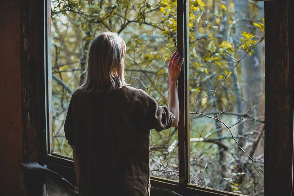person in dark room looking out window after noticing signs of self-harm